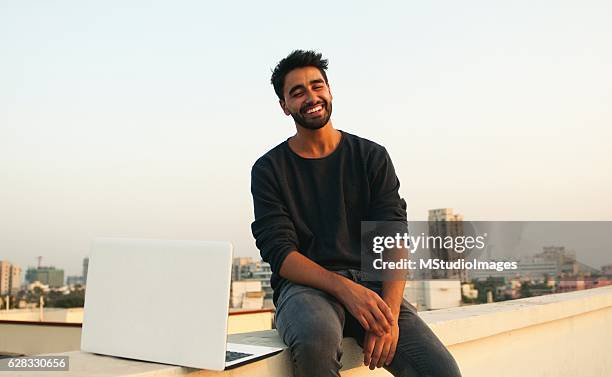 smiling man with laptop on rooftop. - indian ethnicity laptop stock pictures, royalty-free photos & images