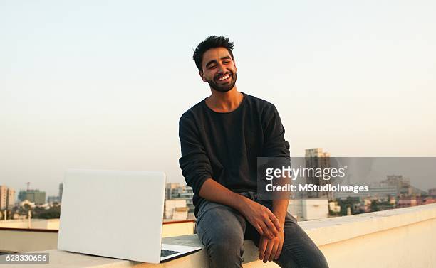 smiling man with laptop on rooftop. - asian man sitting casual imagens e fotografias de stock