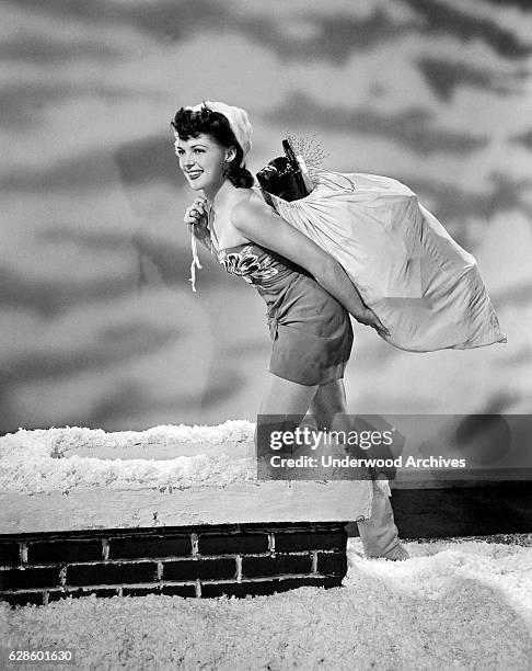 Actress Anne Gwynne is dressed as Santa Claus carrying a bag of gifts as she steps down a chimney, Hollywood, California, circa 1942.