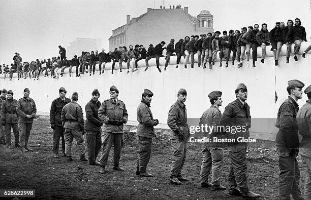 West Berliners line the Berlin Wall in 1989 as East German Soldiers still stand guard over an unopened section at Potsdammer Platz. This view is...