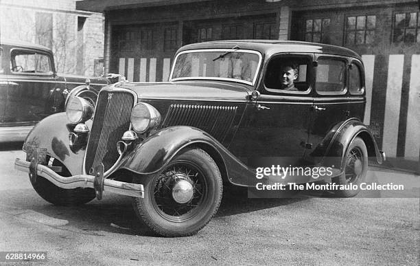 Man sitting in a 1934 Ford DeLuxe Fordor parked in front of garage doors. The 34 Ford is infamous as the car that outlaws Bonnie and Clyde were...