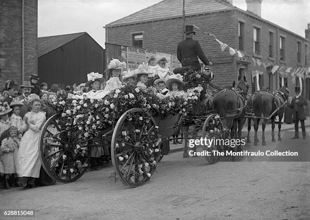 Group of young smartly dressed girls wearing wide brimmed hats and boys in straw tricorn hats sitting in a horse drawn carriage completely covered in...