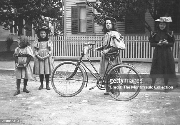 Four young smartly dressed girls standing outside the front of a house, one holding a bicycle with a ribbon tied around the handlebars. Boston...