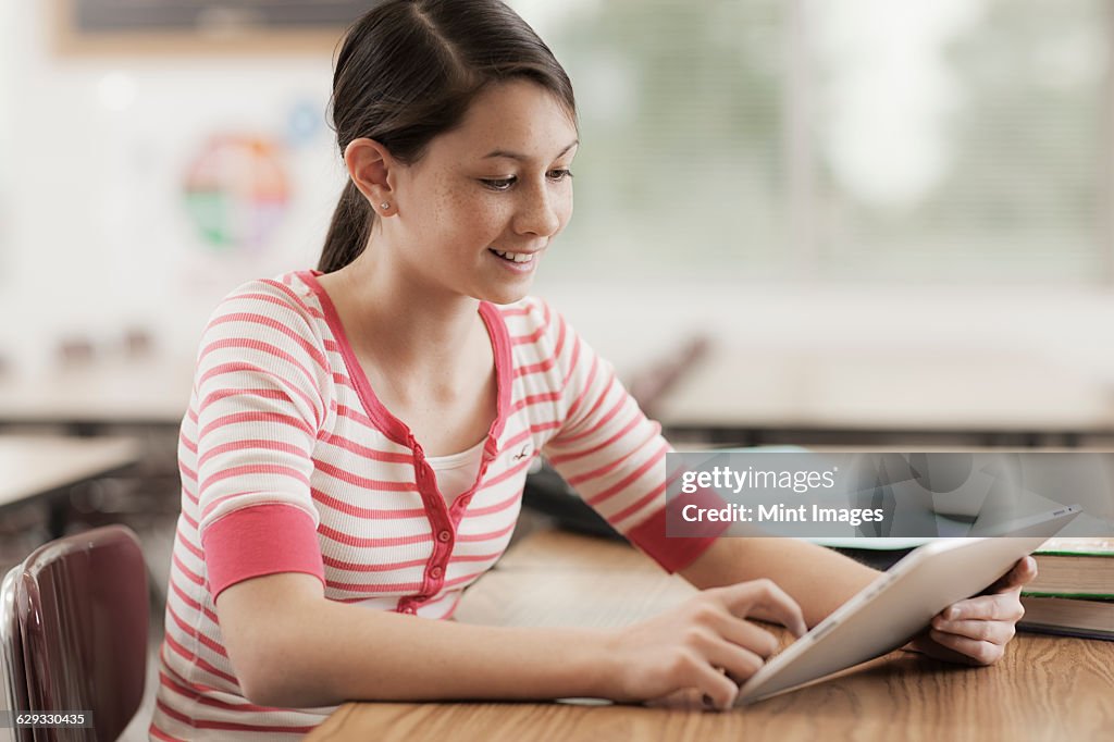 A young girl seated at a desk in a classroom using a digital tablet.