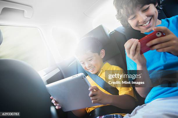 two children travelling in the back seat of a car, one using a digital tablet and one a handheld game. - child car tablet ストッ��クフォトと画像