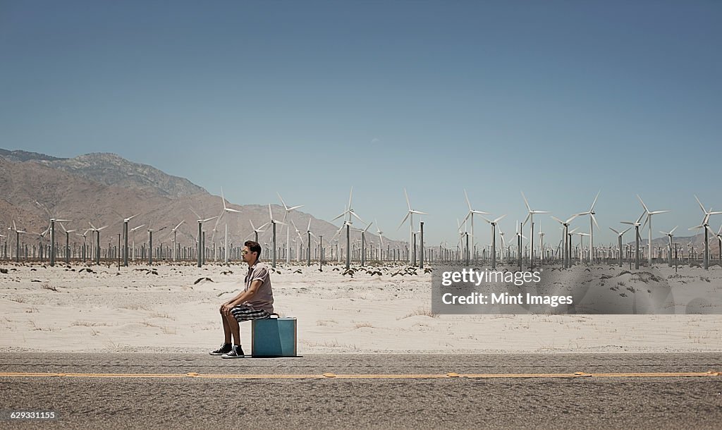 A man standing with a suitcase on the side of a highway, with wind turbines in the background.