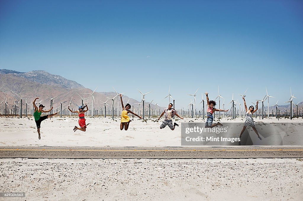 A row of six young people leaping in the air, arms outstretched in wide open space in the desert. 