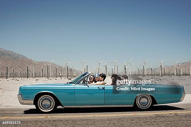 a group of friends in a pale blue convertible on the open road, driving across a dry flat plain surrounded by mountains.  - cabrio stock-fotos und bilder