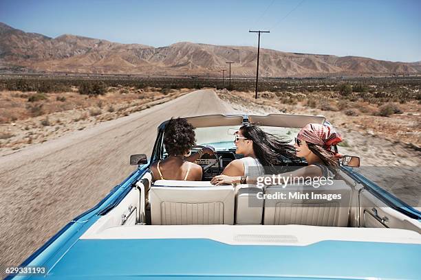 three young people in a pale blue convertible car, driving on the open road across a flat dry plain,  - three people in car stock pictures, royalty-free photos & images