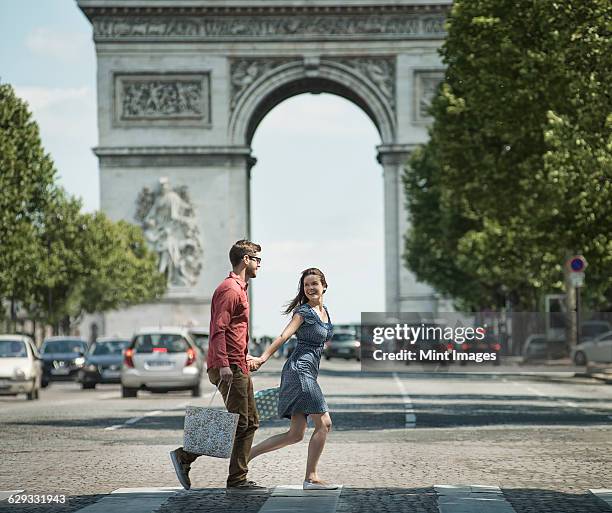 a couple hand in hand carrying shopping bags and crossing the road by a historic monument in the heart of a european city. - foreign photos et images de collection