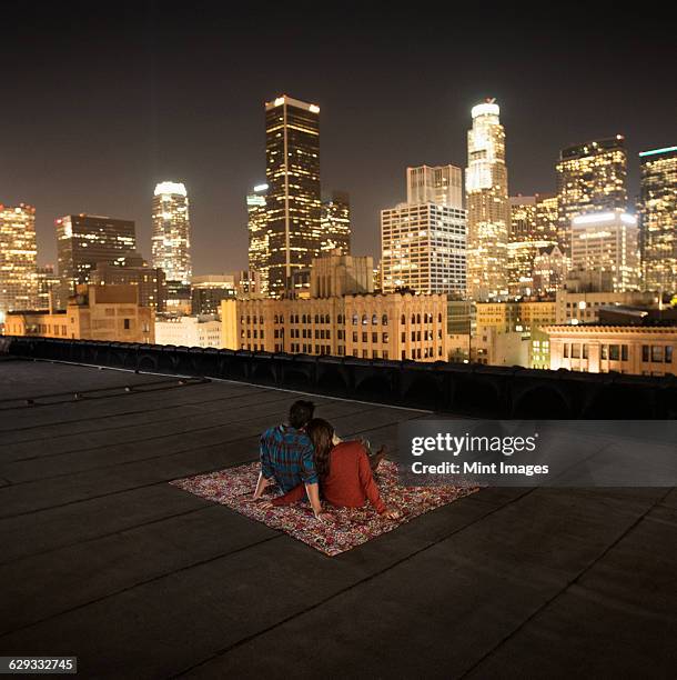 a couple sitting on a rug on a rooftop overlooking a city lit up at night. - rooftop party night stock pictures, royalty-free photos & images