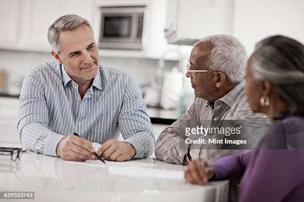three people at a table, a senior couple seated with a man using paper and pen to give them information.  - financial planning seniors stock pictures, royalty-free photos & images