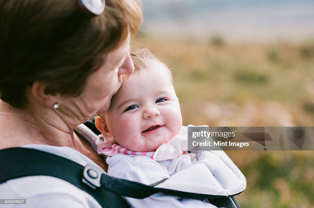 A woman carrying a baby in a baby carrier, kissing her on the head. 