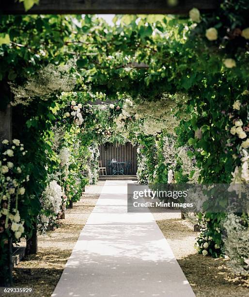 a garden path under a palisade leading to an alcove with table and chairs, with climbing plants and white roses.  - arch foto e immagini stock