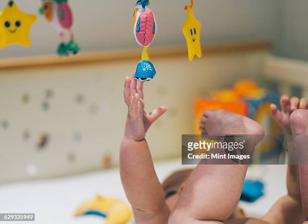 a baby girl in a diaper lying in a cot reaching up to a colourful mobile hanging above her. - móbile - fotografias e filmes do acervo