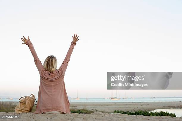 blond woman sitting on a sandy beach, arms raised. - junggesellin stock-fotos und bilder