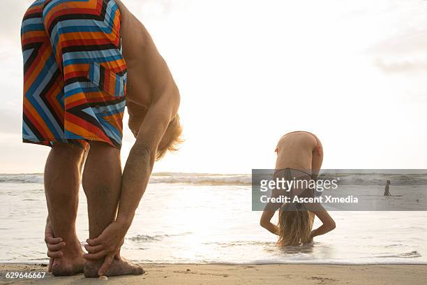 young woman teaches yoga to man on beach - beugen oder biegen stock-fotos und bilder