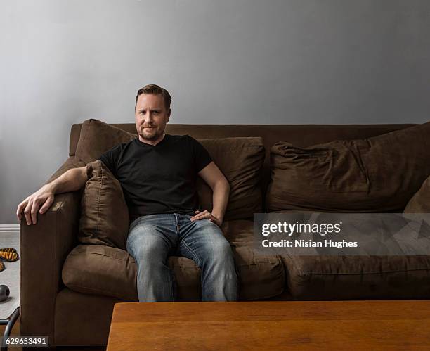 portrait of man sitting on couch looking at camera - man couch foto e immagini stock