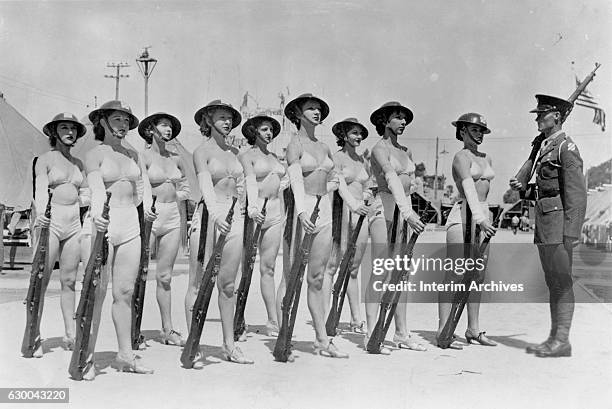 An unidentified corporal from the US Army's 30th Infantry Division stands at attention with a group of women in two-piece bathing costumes, doughboy...