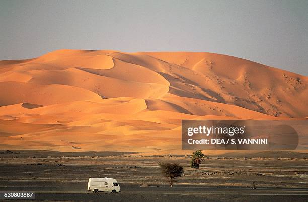 Camper van driving through a desert with dunes, Merzouga, Tafilalt, Ziz valley, Er Rachidia province, Morocco.