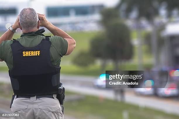 Broward County sheriffs deputy stands on a highway entering Fort Lauderdale International Airport, using binoculars to look towards the airport on...