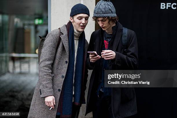 British model Golo Fischer wearing a navy beanie, navy scarf, grey wool coat, skinny black jeans talking and smoking with a model friend during...