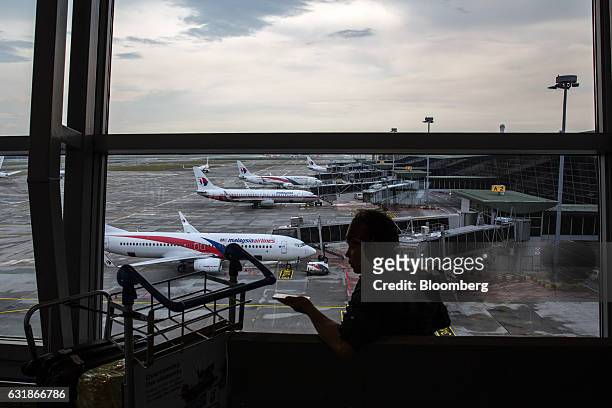 Man uses a smartphone in front of a window while Malaysian Airlines Bhd. Aircraft stand on the tarmac at Kuala Lumpur International Airport in...