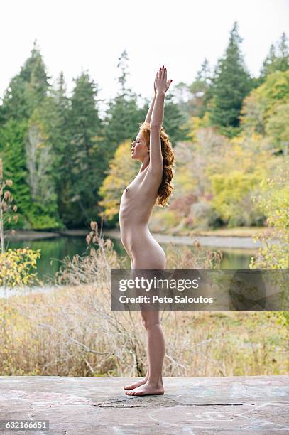 side view of nude young woman meditating on forest lakeside - barefoot redhead ストックフォトと画像