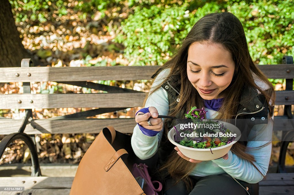 Young woman sitting on park bench eating salad lunch