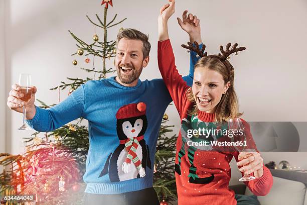 two people with ugly christmas sweaters dancing in front of tree - weihnachtspullover stock-fotos und bilder