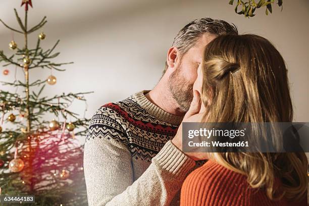 happy couple standing in front of christmas tree, kissing under mistletoe - mistletoe kiss stockfoto's en -beelden