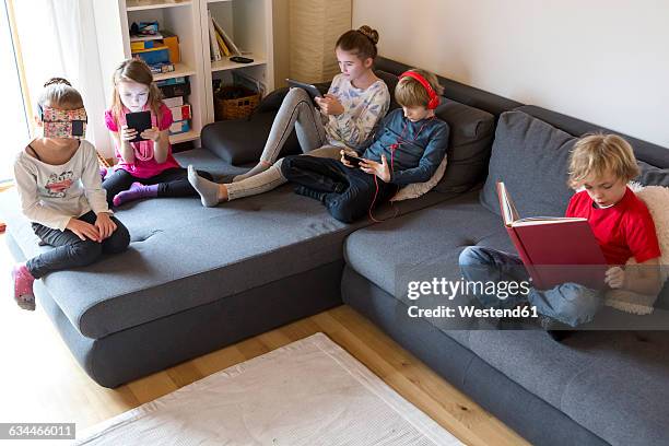 four children on a couch using different digital devices while one boy reading a book - mix media stock pictures, royalty-free photos & images