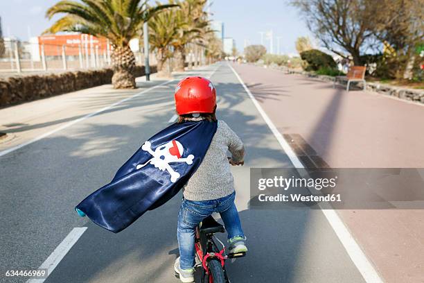 spain, barcelona, back view of little boy with a pirate cape riding bicycle on an empty street - cycling helmet stock pictures, royalty-free photos & images