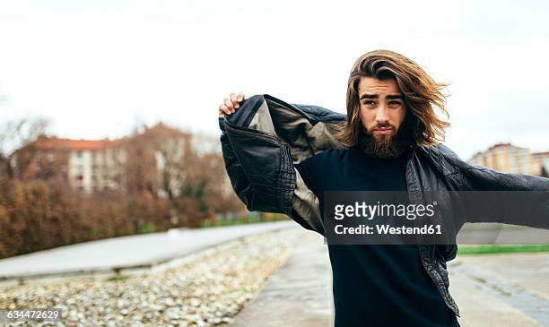 portrait of bearded young man putting on his leather jacket - jacket stock-fotos und bilder