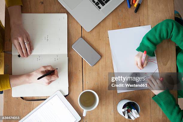 woman at wooden table writing in notebook with son painting a picture - mãe trabalhadora imagens e fotografias de stock