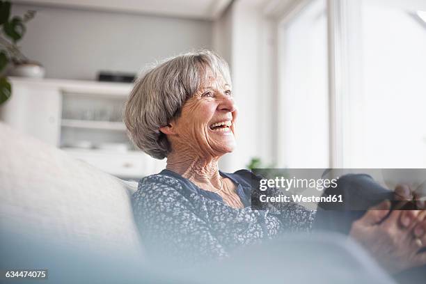 portrait of laughing senior woman sitting on couch at home - nur seniorinnen stock-fotos und bilder