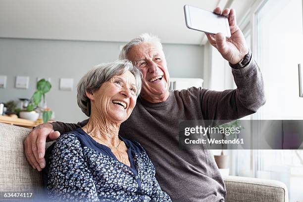 laughing senior couple sitting on the couch in the living room taking selfie with smartphone - joven de espíritu fotografías e imágenes de stock