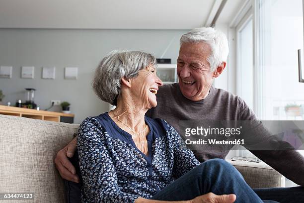 laughing senior couple sitting together on the couch in the living room - coppia anziana foto e immagini stock