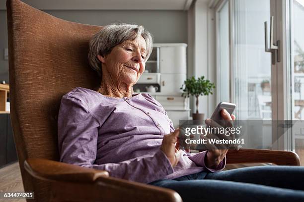 portrait of senior woman sitting on armchair at home using smartphone - sólo mujeres mayores fotografías e imágenes de stock