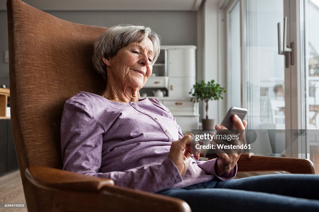 Portrait of senior woman sitting on armchair at home using smartphone