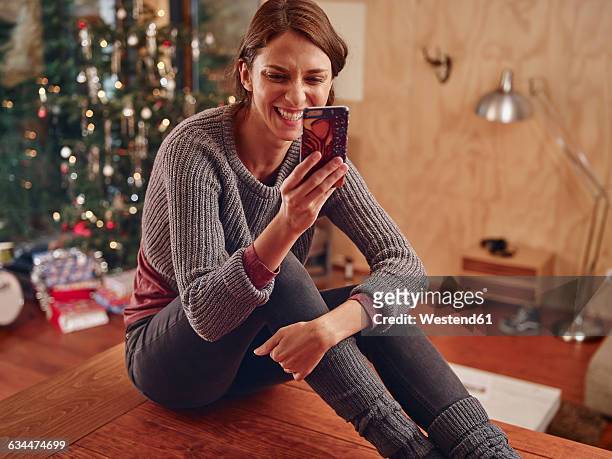 young woman sitting on flooor in front of christmas tree using smart phone - gift lounge stock-fotos und bilder