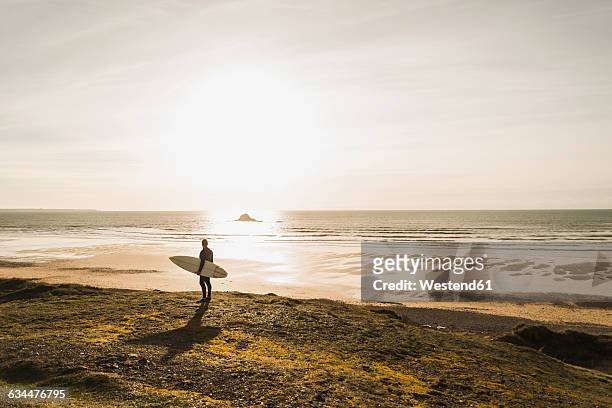 france, bretagne, finistere, crozon peninsula, man standing at the coast at sunset with surfboard - mature surfers stock pictures, royalty-free photos & images