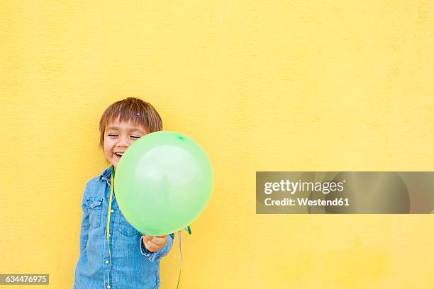 smiling little boy with green balloon and streamer standing in front of yellow wall - child balloon studio photos et images de collection