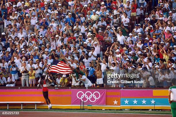 Carl Lewis, running with a large American flag, displays his patriotism and excites the crowd at the 1984 Summer Olympic Games after winning the...
