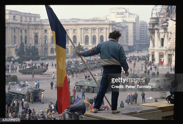 Man holds a Romanian flag with the Communist symbol torn from its center on a balcony overlooking the tanks, soldiers, and citizens filling Palace...