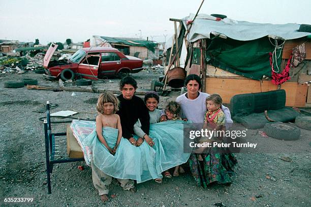 Roma woman and her daughters sit on an old bed near dilapidated shacks and broken down cars. The Roma, or Gypsies, are travelling people and are...