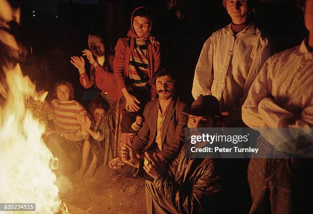 Rom family gathers around a campfire next to a shack at nightfall.