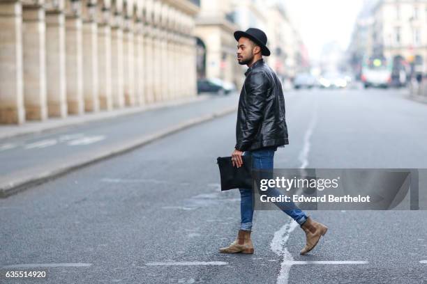 Theo Kimbaloula, fashion blogger, wears a Sand Copenhagen black leather jacket, a Baan Bag black clutch, Asos blue denim jeans, and The Kooples brown...