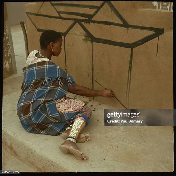 Ndebele woman, with gold hoops covering her lower legs, decorates her house with a design. | Location: Ndebele, Afrique Sud.