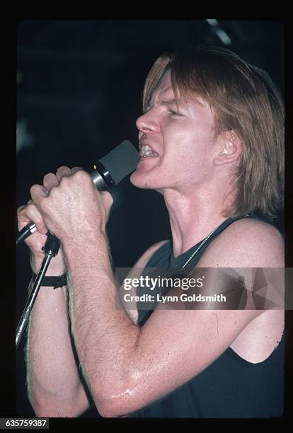 Picture shows writer/singer/poet, Jim Carroll, singing in concert. He is wearing a black tank top and holding a microphone directly in front of his...